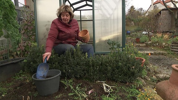 Eine Frau hockt vor einem Gewächshaus und schaufelt Erde.