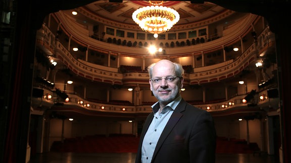 Generalintendant Kay Kuntze im Theatersaal des Landestheaters Altenburg, dunkle, festliche Stimmung, Kronleuchter und Ränge im Hintergrund, im Vordergrund Porträt des Intendanten