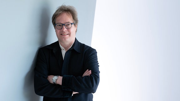 Der Intendant Musikfestspiele Dresden Jan Vogler, ein Mann mit braunem Haar, Brille und schwarzem Anzug, steht mit verschränkten Armen an eine Wand gelehnt. 