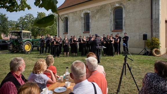 Besucher beim Sommergesang in Diebzig: Blick von einem Biertisch auf einer Wiese in Richtung einer Dorfkirche, vor der ein Traktor steht und ein Chor singt