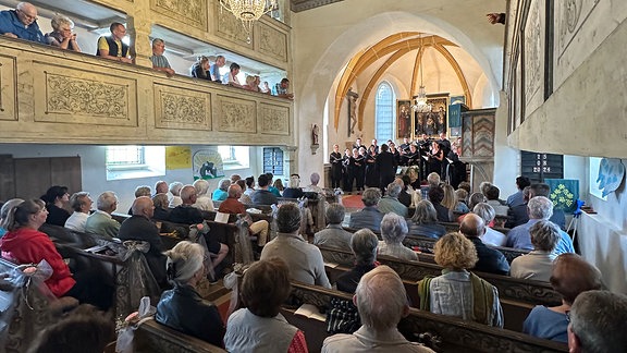 Besucherinnen und Besucher in einer schmuckvollen Kirche. Man sieht die Besuchenden von hinten, sie blicken auf den Altarraum, wo sich der Chor mit Dirigent versammelt hat. 