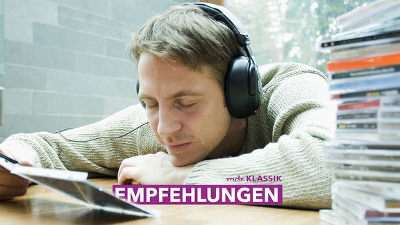 Ein Mann mit Kopfhörern lehnt auf dem Tisch neben einem CD-Stapel, in der Hand eine offene CD-Hülle.