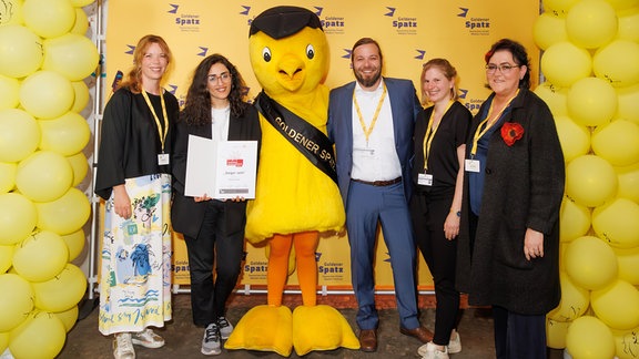 Vier Frauen und ein Mann stehen mit einer als gelber Vogel kostümierten Figur vor eine gelben Wand mit gelben Luftballons.