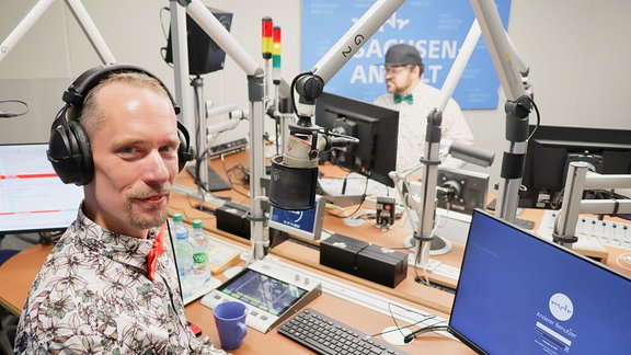 Zwei Männer mit Hemd und Fliege stehen an Mikrofonen in einem Radio-Studio