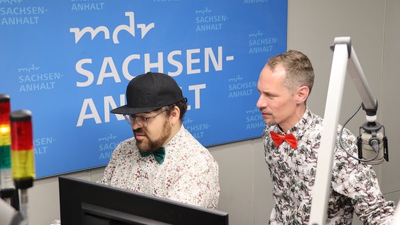 Zwei Männer mit Hemd und Fliege stehen an Mikrofonen in einem Radio-Studio