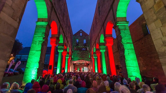 Das Kulturfestival im Kloster Paulinzella: Die Mauern des Klosters sind grün und rot angestrahlt. Dazwischen sitzt Publikum. 