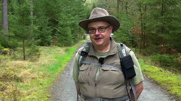 Ein Mann steht auf einem Waldweg und schaut in die Kamera.