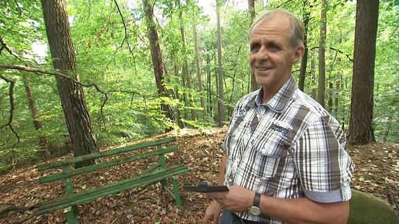 Ein Mann steht neben einer Sitzbank im Wald und lächelt.