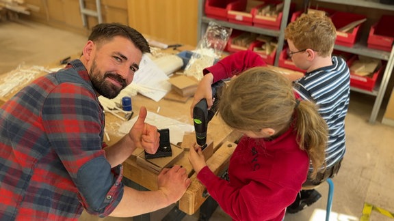 Tischler zeigt Daumen hoch während er mit zwei handwerkelnden Kindern an einem Tisch sitzt