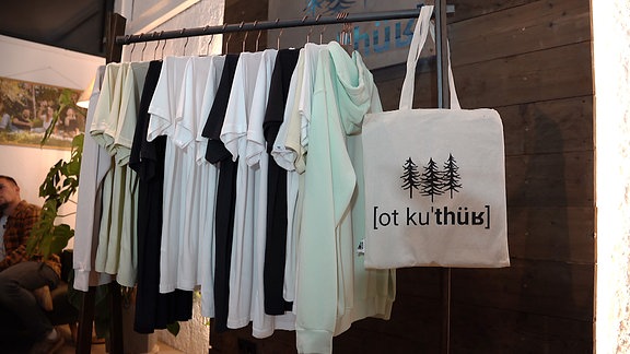 T-Shirts und Pullis, sowie ein Beutel mit der Aufschrift "ot ku'thür hängen in einem Geschäft an einer Kleiderstange.