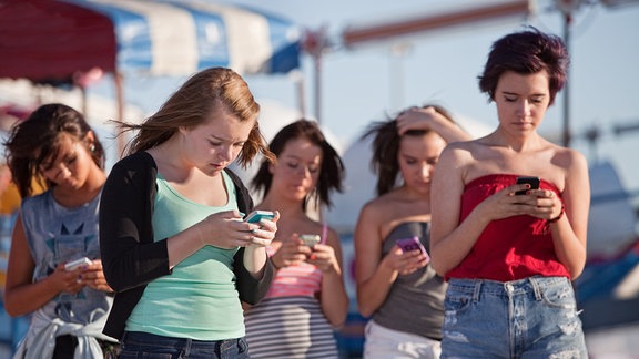 Mehrere junge Frauen stehen im Sonnenschein und blicken jeweils auf ihr eigenes Smartphone. 