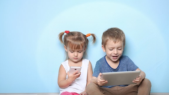 Zwei Kleinkinder sitzen nebeneinander und haben ein Smartphone und ein Tablet in der Hand.