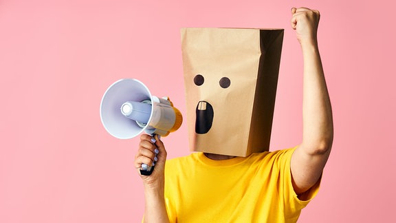 Person mit Papiertüte auf dem Kopf schreit in ein Megafon.