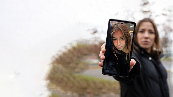 Eine Frau hält einen Smartphonebildschirm in die Kamera, auf dem ein junges Mädchen abgebildet ist.