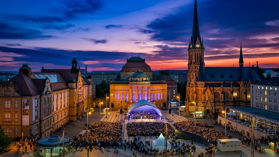 Theaterplatz in Chemnitz im Abendlicht, darauf eine erleuchtete Bühne und Publikum