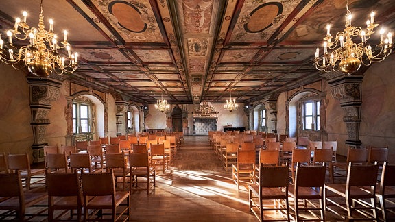 Blick durch zwei Stuhlreihen in einen großen Saal mit Kronleuchtern und einer reich verzierten Decke