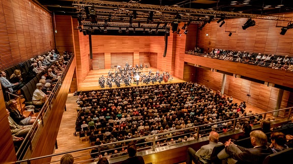 Blick über die Konzertgäste auf die Bühne der Weimarhalle in Weimar, darauf ein Orchester