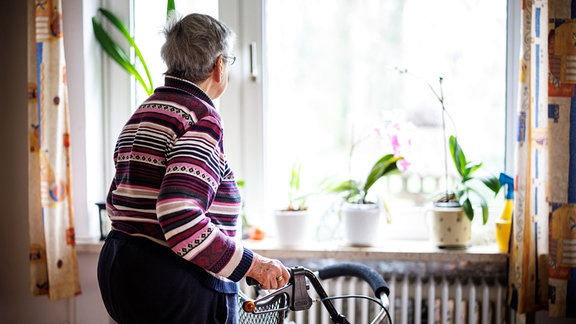 Eine ältere Dame blickt aus einem Fenster ihrer Wohnung und stützt sich dabei auf ihrem Rollator ab.