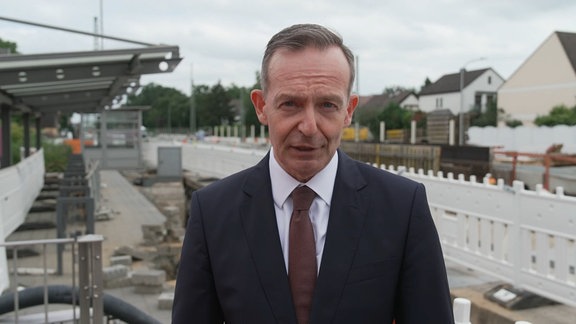 Bundesverkehrsminister Volker Wissing blickt beim Interview in die Kamera. Hinter ihm eine Baustelle der Bahn.