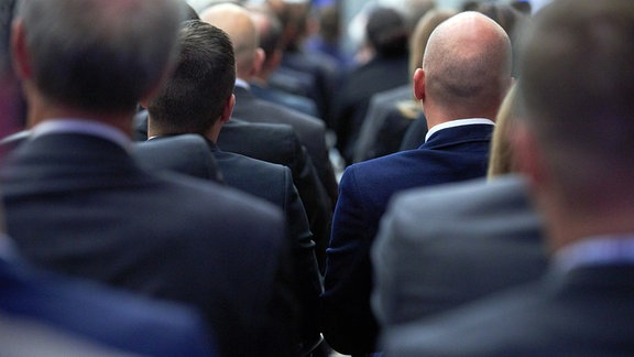 Männer in dunklen Anzügen sitzen in mehereren Reihen nebeneinander.