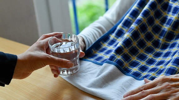 Ein*e Pfleger*in reicht einer alten Frau ein Glas Wasser. 