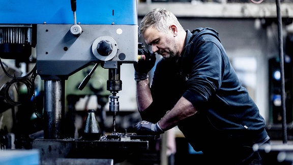 Älterer Mann steht in blauer Trainingsjacke an einer Maschine und bohrt ein Loch in eine Metallplatte 