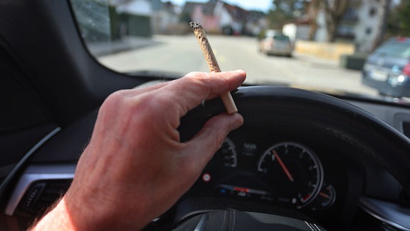 Ein Mann sitzt mit einem Joint zwischen den Fingern am Steuer eines Autos. 