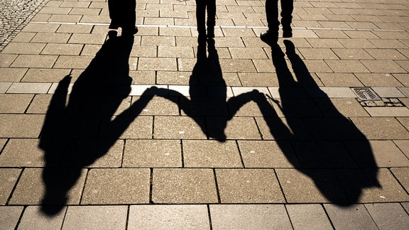 Die Schatten von zwei Erwachsenen und einem Kind sind in den Morgenstunden auf dem Asphalt in der Innenstadt zu sehen. 