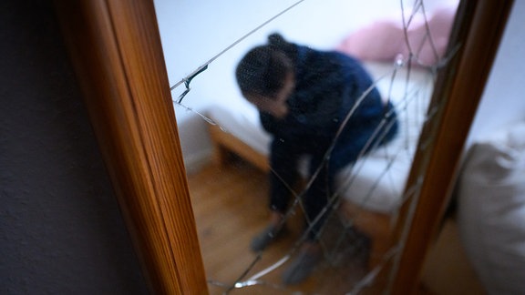 Eine Frau sitzt neben einem gesprungenen Spiegel auf einem Bett und hält ihren Kopf in den Händen (gestellte Szene).