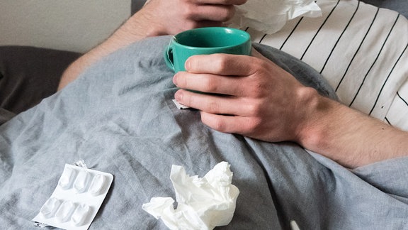 Ein Mann liegt mit Taschentüchern, Teetasse, Nasenspray und Tabletten im Bett (gestellte Szene).