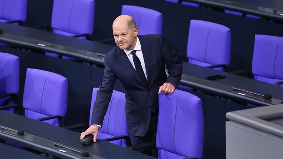 Bundeskanzler Olaf Scholz (SPD) hält sich an einem Stuhl auf der Regierungsbank fest und legt etwas ab.