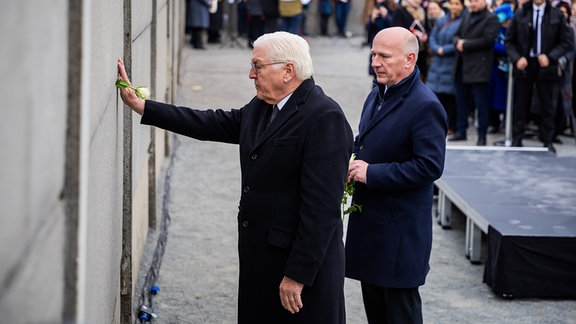 Bundespräsident Frank-Walter Steinmeier (l) steckt neben Kai Wegner (CDU), Regierender Bürgermeister von Berlin, bei einer zentralen Gedenkveranstaltung anlässlich von 35 Jahre friedliche Revolution und Mauerfall in der Gedenkstätte Berliner Mauer eine Rose in die Hinterlandmauer. 