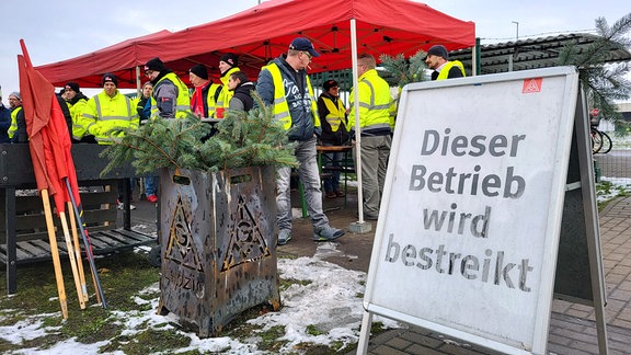 Mitarbeiterinnen und Mitarbeiter der Schrottrecyclingfirma SRW metalfloat Espenhain streiken in neongelben Warnwesten unter einem roten Zelt neben einer Feuertonne der IG Metall Leipzig, im Vordergrund steht ein Aufsteller mit der Aufschrift "Dieser Betrieb wird bestreikt"