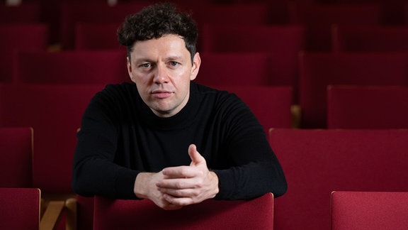 Der deutsche Schauspieler und Sänger Christian Friedel sitzt in den Sitzreihen eines Theaters und blickt ernst in die Kamera