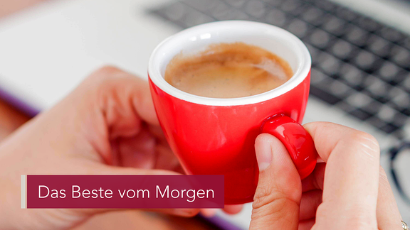 Jemand hält eine rote Espresso-Tasse in der Hand. Der Kaffee dampft. Auf einem Tisch liegen ein Laptop und eine Brille.