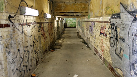 Blick in einen maroden Personentunnel, an den Wänden Graffitis, auf dem Boden Dreck und Wasserflecken.