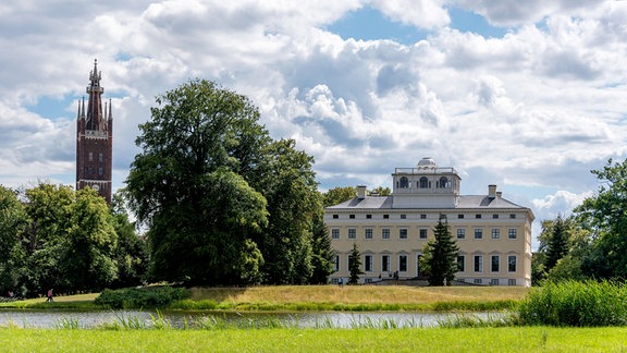 Blick auf ein herrschaftliches, von Bäumen umgebenes Gebäude, im Vordergrund ein Fluss