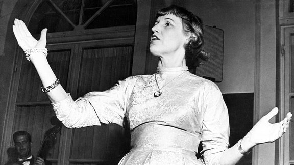 Lotte Lenya, in einem eleganten Kleid, steht singend mit ausgebreiteten Armen in einem Raum. Rings herum sitzen mehrere Männer.