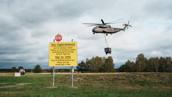 Ein Bundeswehr-Hubschrauber hebt mit angehängter Last ab. Im Vordergrund steht ein Schild, das vor dem Flugbetrieb warnt.