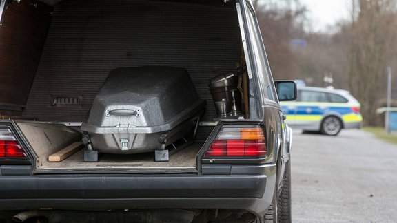 Leichenwagen mit Sarg auf der Ladefläche auf einem Weg vor einem Polizeiauto