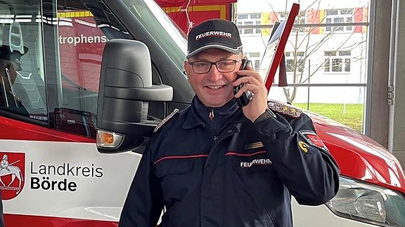 Ein Mann steht mit Handy am Ohr in Feuerwehr-Kleidung vor einem Rettungswagen