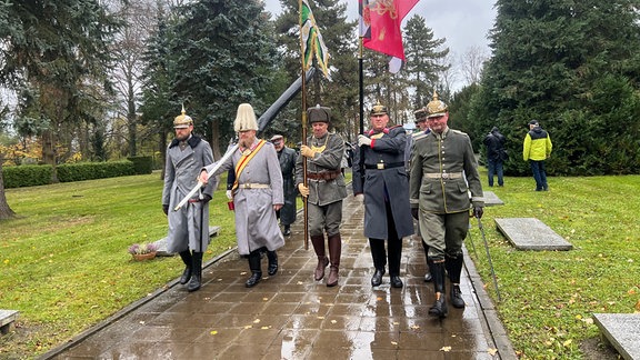 Männer in historischen Militäruniformen laufen auf einem Friedhof. 