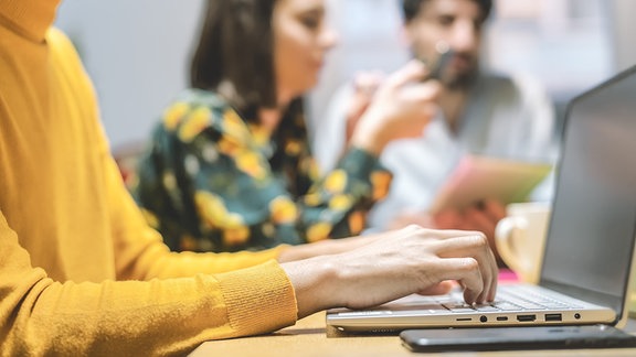 Drei junge Personen sitzen an einem Tisch und arebeiten an Laptop oder Smartphone.