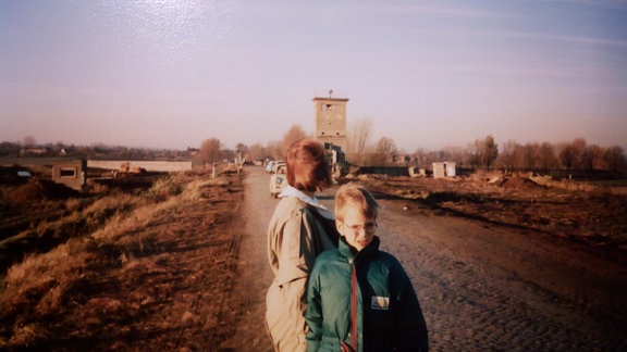 Eine Frau und ihr Sohn stehen auf einer Landstraße vor einem Wachturm. 