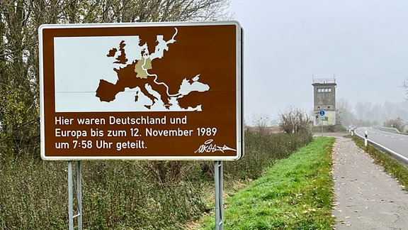 Ein braunes Gedenkschild steht neben einer Straße, die zu einem alten Wachturm führt.