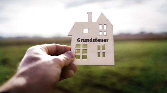 Hand hält ein Haus aus Holz mit der Aufschrift: Grundsteuer