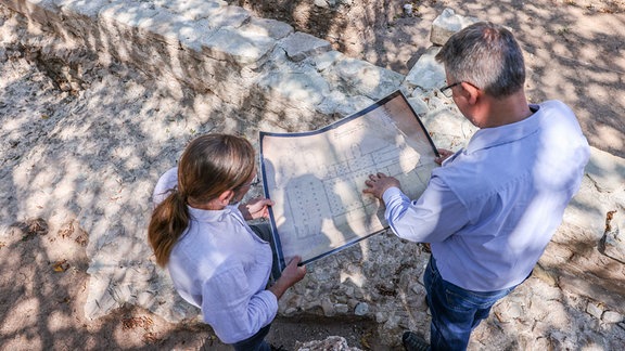 Zwe  Projektleiter, stehen mit einem historischen Plan bei Grabungsarbeiten auf dem Areal des ehemaligen Klostergeländes Posa