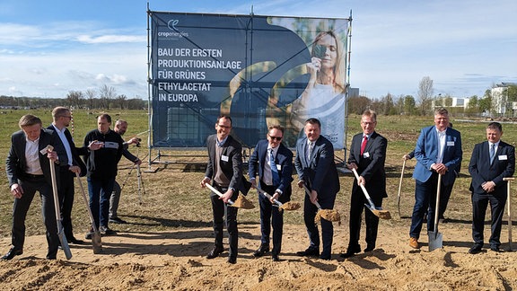 Viele Männer mit Anzug und Spaten stehen auf einem Feld.