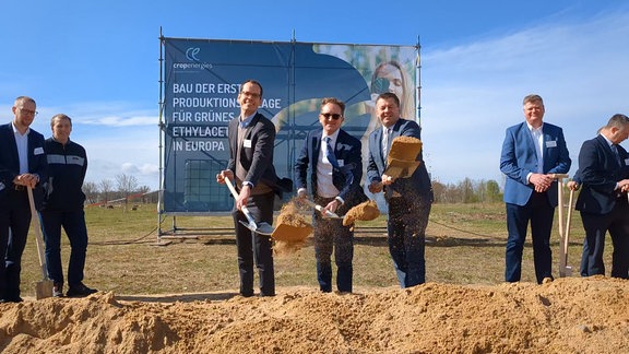 Drei Männer mit Anzug und Spaten schippen Sand.