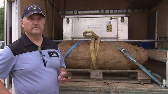 Ein Mann vom Kampfmittelbeseitigungsdienst mit einem Zünder in der Hand vor einer alten Bombe